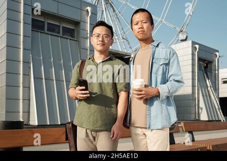 Zwei junge Geschäftsleute in Casualwear blicken auf Sie, während sie vor der Kamera gegen moderne Architektur und Vergnügungspark stehen Stockfoto