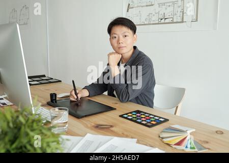 Junge ernsthafte Designerin mit Stylus-Stift Zeichnung Skizze auf dem Bildschirm des grafischen Tabletts, während Sie am Arbeitsplatz sitzen Stockfoto