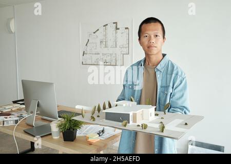 Der junge, selbstbewusste Architekt hält das Layout des Hauses, während er vor der Kamera gegen seinen Arbeitsplatz im Büro steht Stockfoto