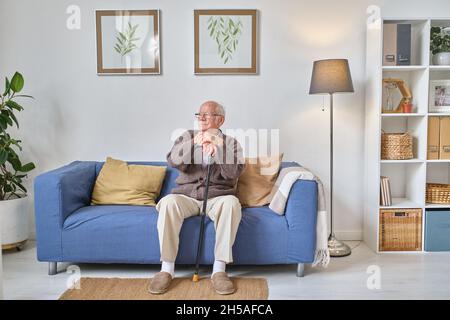 Älterer Mann mit Krücke sitzt und ruht sich auf dem Sofa im Wohnzimmer aus Stockfoto