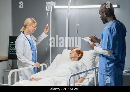 Arzt verschreibt Medikamente, während die Krankenschwester einen Tropf herstellte, behandelte sie ältere Frauen auf der Krankenhausstation Stockfoto