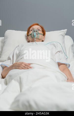 Ältere Frau in Sauerstoffmaske, die während des Coronavirus auf dem Bett liegt und mit speziellen medizinischen Geräten auf der Krankenhausstation atmet Stockfoto