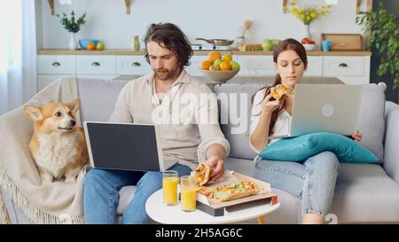 Junges Paar, das zusammen auf der Couch sitzt, eine Pizza isst und mit einem Laptop arbeitet. In der Nähe von Sitting A Cute Dog. Stockfoto