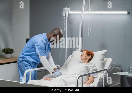 Afrikanischer männlicher Arzt in Uniform, der ältere Frau, die im Krankenhaus auf dem Bett liegt, tropfer macht Stockfoto