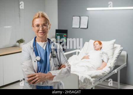 Porträt einer reifen Ärztin im weißen Mantel, lächelnd in der Kamera stehend, mit gekreuzten Armen und einem kranken Patienten, der im Hintergrund im Bett liegt Stockfoto