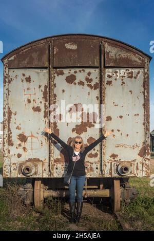 Erwachsene Frau mit Sonnenbrille, lächelnd, mit erhobenen Armen, gelehnt an einen verlassenen Zugwagen. Stockfoto