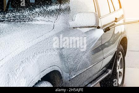 Autowäsche mit Active-Foam-Shampoo im Sommer im Freien in der Autowaschstation. Das Äußere des Fahrzeugs ist mit einer Schicht aus weichem Schaum und Schaumsprühen aus dem Schlauch bedeckt. Stockfoto