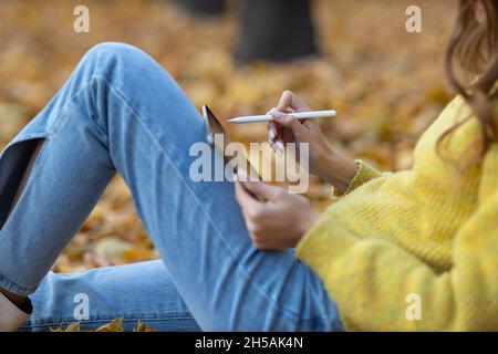 Junge Frau Modell sitzt mit digitalen Tablet und malen mit Bleistift Stift im Herbst Park mit gelbem Laub Ahornblätter. Stockfoto