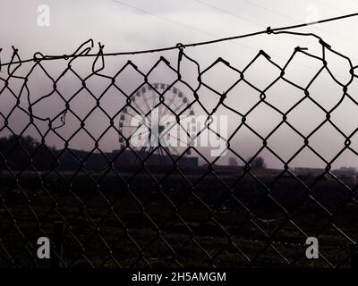 Sicherheitszaun Stacheldraht mit großem Feld und Karussell-Riesenrad lustig gehen im Hintergrund durch Nebel gesehen Stockfoto