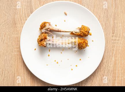 Zwei Hühnerknochen, die auf einem weißen Teller übrig sind. Ungesunde Fast Food, Hühnerbeine, Draufsicht Stockfoto