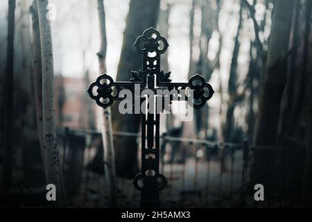 In der beängstigenden Dämmerung steht auf einem verlassenen Friedhof zwischen den Bäumen ein schwarzes altes katholisches Kreuz. Horror und Angst. Religion und Tod. Stockfoto