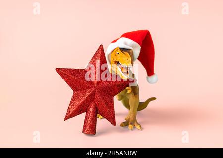 Dinosaurier Rex in rot Weihnachtsmann Hut hält Stern in den Pfoten auf rosa Hintergrund Silvester oder Heiligabend Art Weihnachtskarte Kreative Idee für Merry Stockfoto