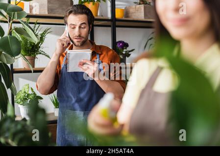 Junge Floristin, die am Handy sprach und Notebook in der Nähe von Pflanzen ansah, und verschwommener Kollege im Geschäft Stockfoto