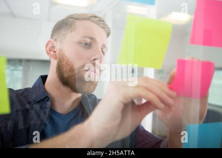 Nachdenklicher Geschäftsmann, der sich Haftnotizen auf dem Fenster ansieht Stockfoto