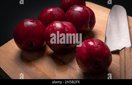 Rote Pflaumen auf Brotbrett mit Messer und schwarzem Hintergrund Stockfoto