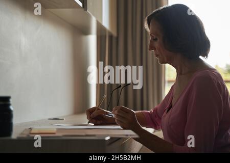 Eine ältere Dame, die am Schreibtisch saß und auf Papier schrieb Stockfoto