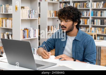Junger indischer Student, der sich das Webinar mit einem Laptop in der Bibliothek ansieht Stockfoto