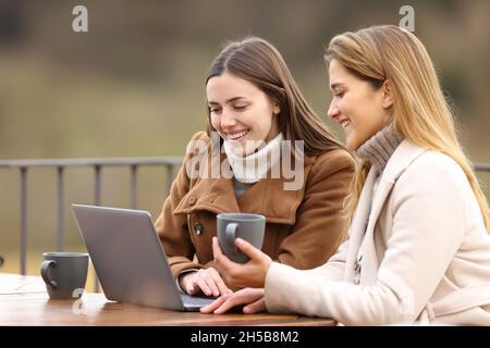 Zwei glückliche Freunde, die im Winter ihren Laptop checken und Kaffee trinken, sitzen auf einer Terrasse Stockfoto