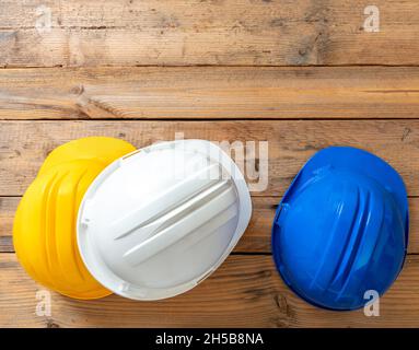 Schutzhelme, weiße blaue und gelbe Schutzhelme. Baustellenarbeit persönliche Schutzkopfausrüstung, Holzhintergrund. Harthüte für Ingenieure Stockfoto
