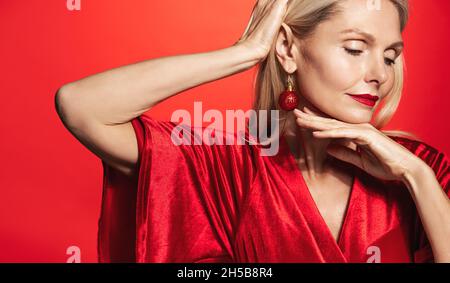 Elegante 50 Jahre alte Frau, elegante reife Dame mit Weihnachtsohrringen und rotem Kleid, Urlaub Make-up, berühren ihr Gesicht, zeigt glühende gesunde Haut Stockfoto