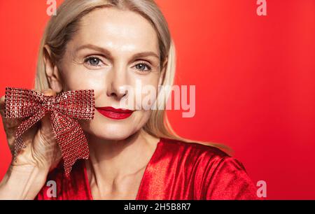 Lächelnd elegante Frau 50 Jahre alt, reife Dame mit Überraschungsgeschenk Band, weihnachtsgeschenk in der Hand, trägt roten Lippenstift und Abend festlich Stockfoto