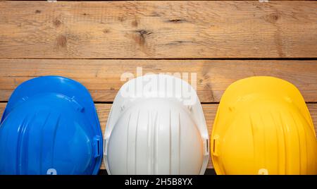 Schutzhelme, weiße blaue und gelbe Schutzhelme. Baustellenarbeit persönliche Schutzkopfausrüstung, Holzhintergrund. Harthüte für Ingenieure Stockfoto