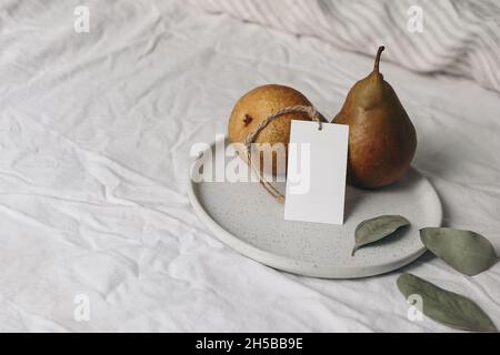 Gelbe Birnen Früchte auf Keramikplatte Blank Geschenkanhänger Etikett Mockup mit trockenen Eukalyptusblättern. Herbst, Sommer Verkauf Zusammensetzung.Weiß gestreiftes Leinen Stockfoto