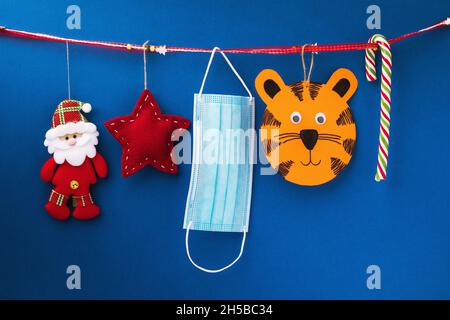 2022 Symbol, weihnachtsbaumspielzeug und medizinische Einwegmaske hängen auf dekorativem Band auf blauem Hintergrund, weihnachts- und Neujahr-Quarantänekonzept Stockfoto