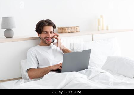Fröhlicher attraktiver Tausendjähriger europäischer Mann arbeitet am Laptop, telefoniert mit dem Kunden im Bett Stockfoto
