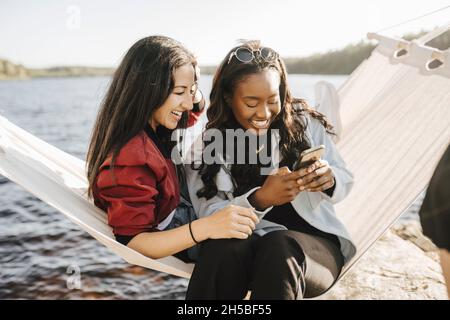 Glückliche weibliche Freunde, die Smartphone benutzen, während sie auf Hängematte sitzen Stockfoto