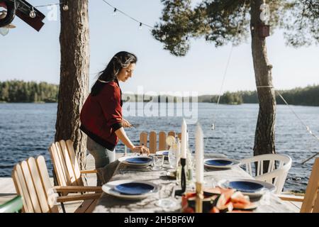 Junge Frau, die am sonnigen Tag am See Tisch legt Stockfoto