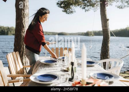 Lächelnde junge Frau, die an einem sonnigen Tag am See Tisch legt Stockfoto
