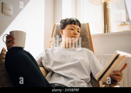 Junge Frau liest Buch, während sie zu Hause die Kaffeetasse hält Stockfoto