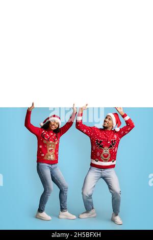 Fröhliche schwarze Liebhaber in Weihnachtsmützen, die unter leerem Plakat stehen Stockfoto