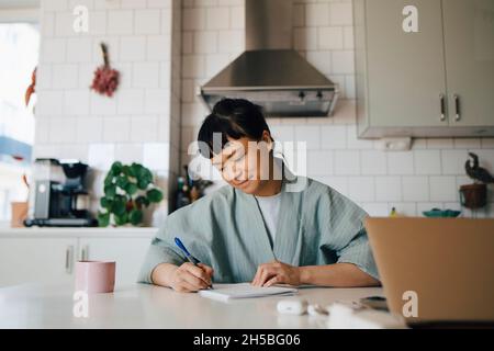 Junge Frau im Bademantel, die in der heimischen Küche im Buch schreibt Stockfoto