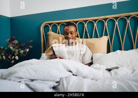 Lächelnder Mann mit mittlerem Erwachsenenalter, der zu Hause im Bett ein Buch liest Stockfoto