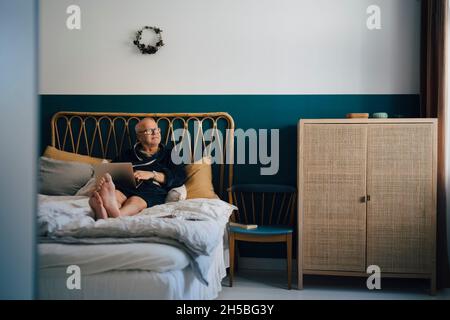 Ein älterer Mann blickt weg, während er zu Hause mit einem Laptop auf dem Bett sitzt Stockfoto