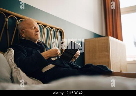 Älterer Mann mit geschlossenen Augen hält Buch, während er auf dem Bett sitzt Stockfoto