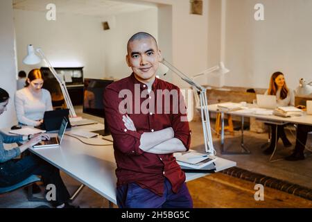 Selbstbewusster männlicher Unternehmer, der sich mit gekreuzten Armen am Schreibtisch im Büro lehnt Stockfoto