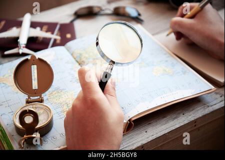 Männliche Hand hält eine Lupe für die Planung Reise mit Weltkarte auf dem Tisch. Reisekonzept. Sonnenbrille, Pässe, Notizbuch, Kompass, ca. Stockfoto