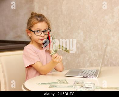 Mädchen 5 Jahre alt in Brille, mit Dollar-Rechnungen und ein Computer spielt Buchhalter. Konzept der Remote-Arbeit in Quarantäne, Kinderentwicklung. Vertuelles Spiel. Stockfoto