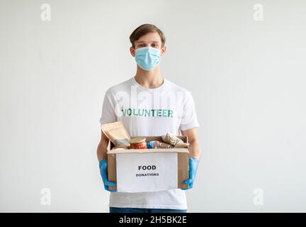 Porträt eines jungen Mannes mit T-Shirt, medizinischer Gesichtsmaske und Handschuhen, der Spendenbox über der hellen Wand hält Stockfoto