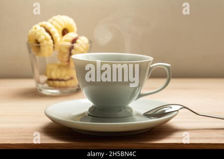 Eine weiße Tasse mit heißem Kaffee, der ausdampft, und ein kleiner Glasaufsatz mit Keksen auf einer Holzoberfläche. Stockfoto