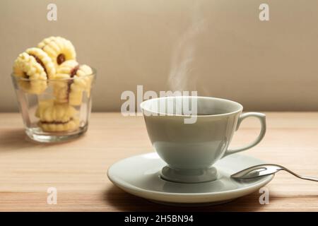 Eine weiße Tasse mit heißem Kaffee, der ausdampft, und ein kleiner Glasaufsatz mit Keksen auf einer Holzoberfläche. Stockfoto