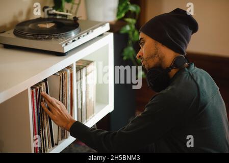 Junger Tausendjähriger Mann mit gemischtem Rennen in Beanie mit Kopfhörern, der sich in der Vinylsammlung umsah und dabei war, Musik zu hören. Stockfoto