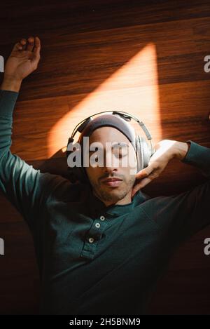 Ein junger Mann mit gemischter Rasse, der auf dem Boden liegt und Musik im Sonnenlicht mit Kopfhörern hört. Hübscher Hipster, der auf dem Rücken liegt und sich zur Musik entspannt Stockfoto