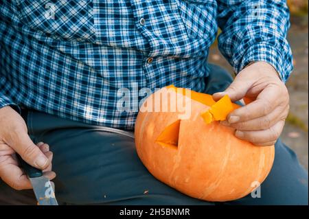 Ein alter Mann schneidet Löcher in einen Kürbis. Ein Mann sitzt auf einer Bank im Garten und kocht einen Kürbis für die Halloween-Feiertage. Stockfoto