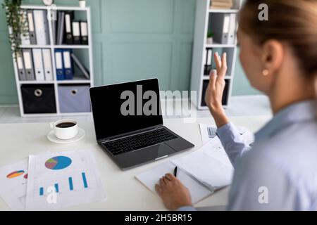 Remote-Meeting. Über die Schulter blicken Sie auf eine Geschäftsfrau, die am Schreibtisch sitzt und über einen Laptop spricht und die Webcam anwinkt Stockfoto