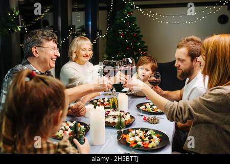 Lebhafte große Familie bei der weihnachtsfeier. Sitzen um einen großen Esstisch, klirren Weingläser, feiern. Weihnachtsbaum und Girlanden in Backgr Stockfoto