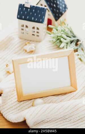Winterurlaub Stillleben. Fotorahmen-Modell auf gestricktem karierten Tannenzweigen, beigefarbenen Kugeln, Dekoration kleiner Häuser Stockfoto